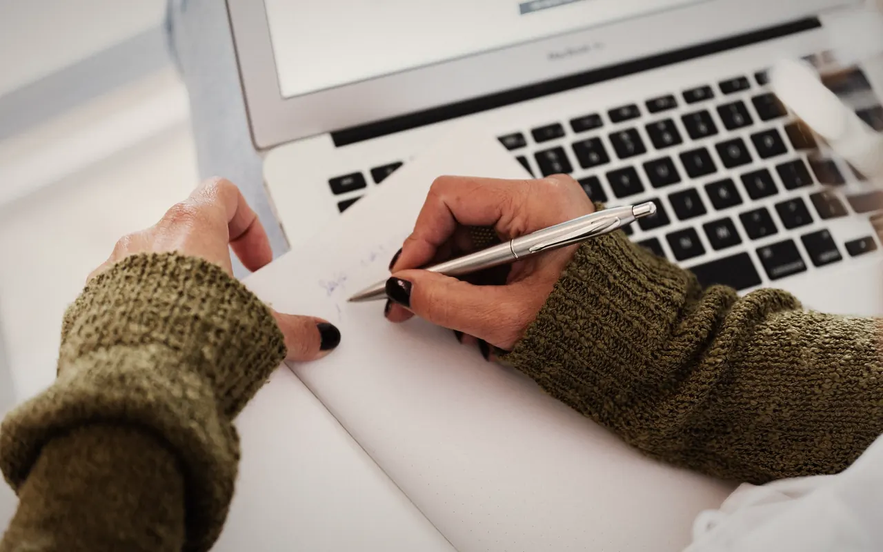 Person writing down trades in a notebook next to a laptop showing a chart